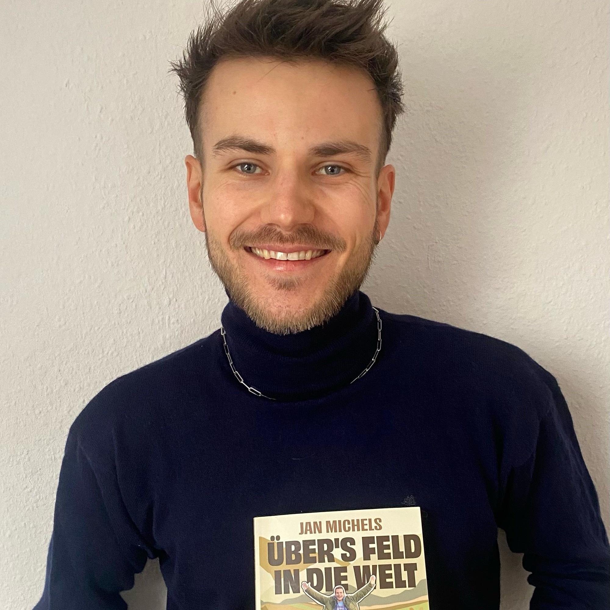 Jan Michels mit seinem Buch „Übers Feld in die Welt“. Er trägt einen dunkelblauen Rollkragenpullover mit silberner Kette.
