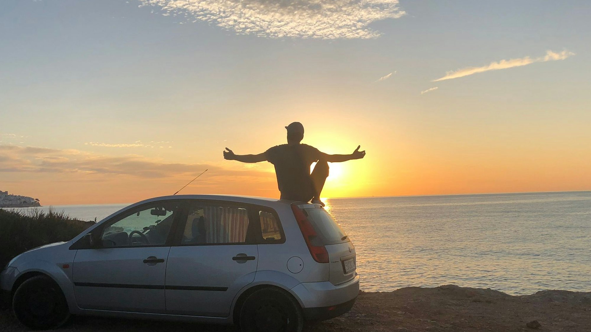 Ein Mann sitzt mit ausgebreiteten Armen auf dem Dach eines älteren Ford Fiesta, der auf einer Anhöhe über dem Meer steht. Über dem Meer geht die Sonne unter.
