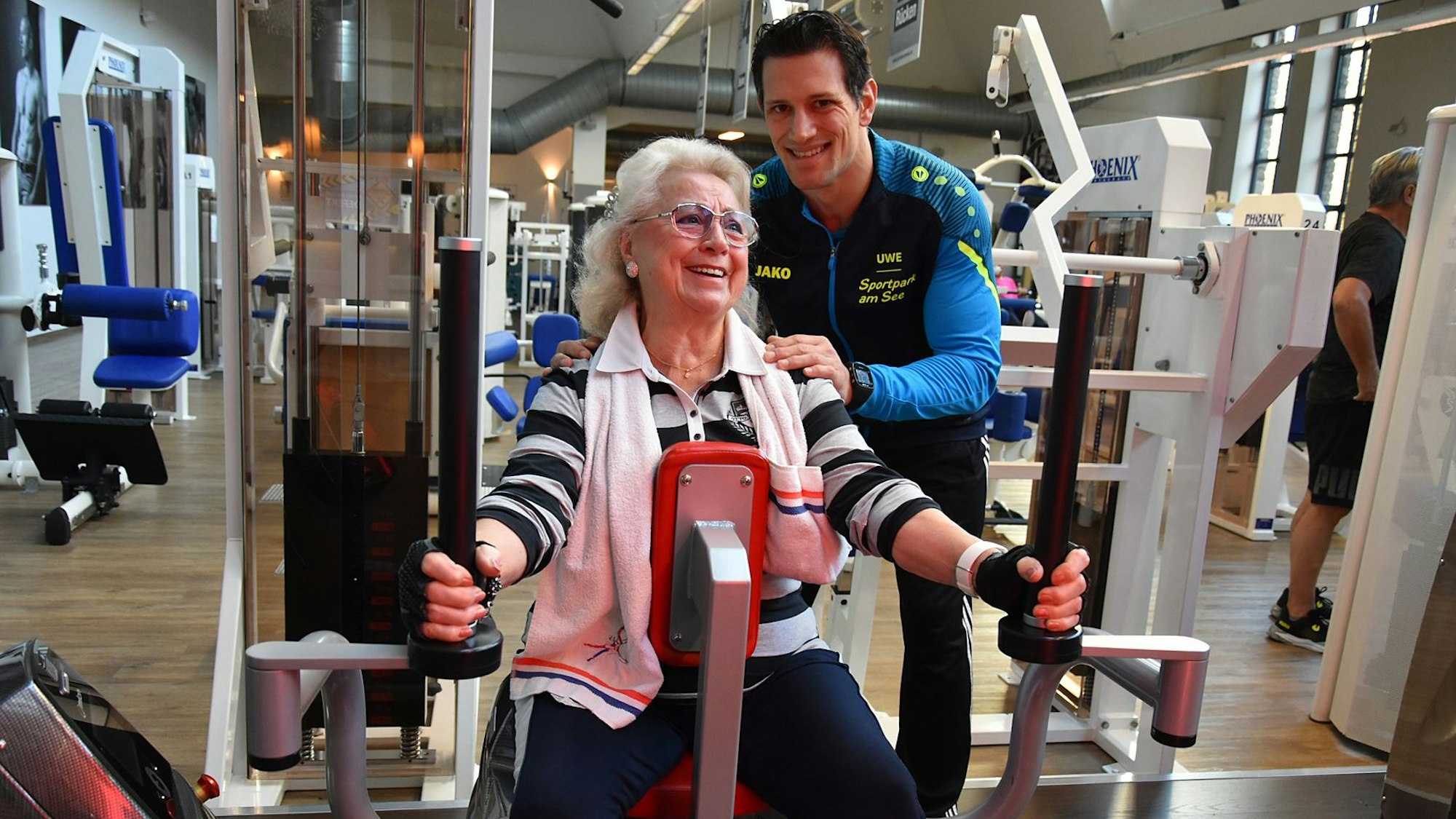 Eine ältere Dame am Kraftgerät, dahinter ein Trainer.