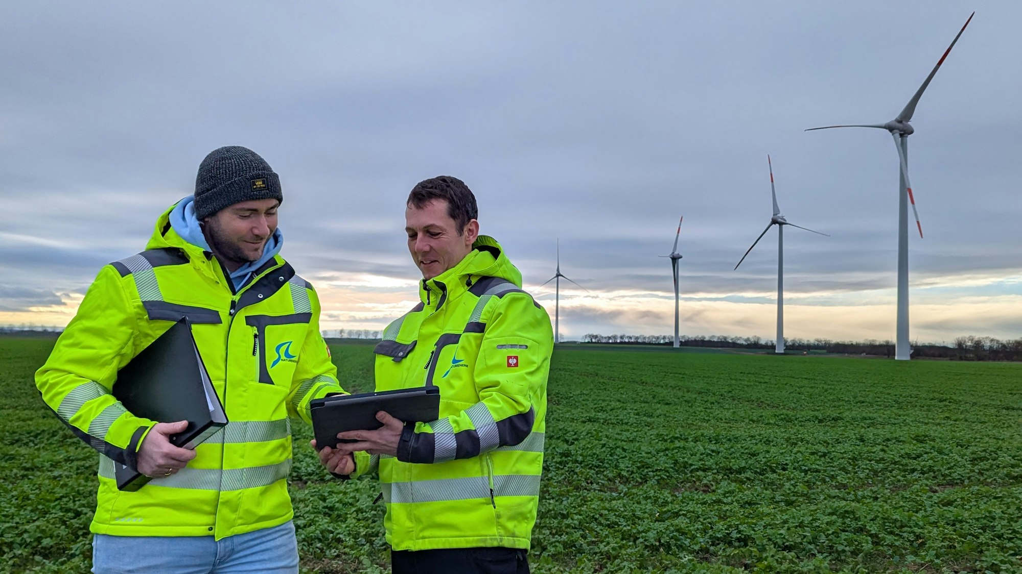 Das Foto zeigt zwei Männer in leuchtenden Warnjacken, dahinter die vier Windräder.