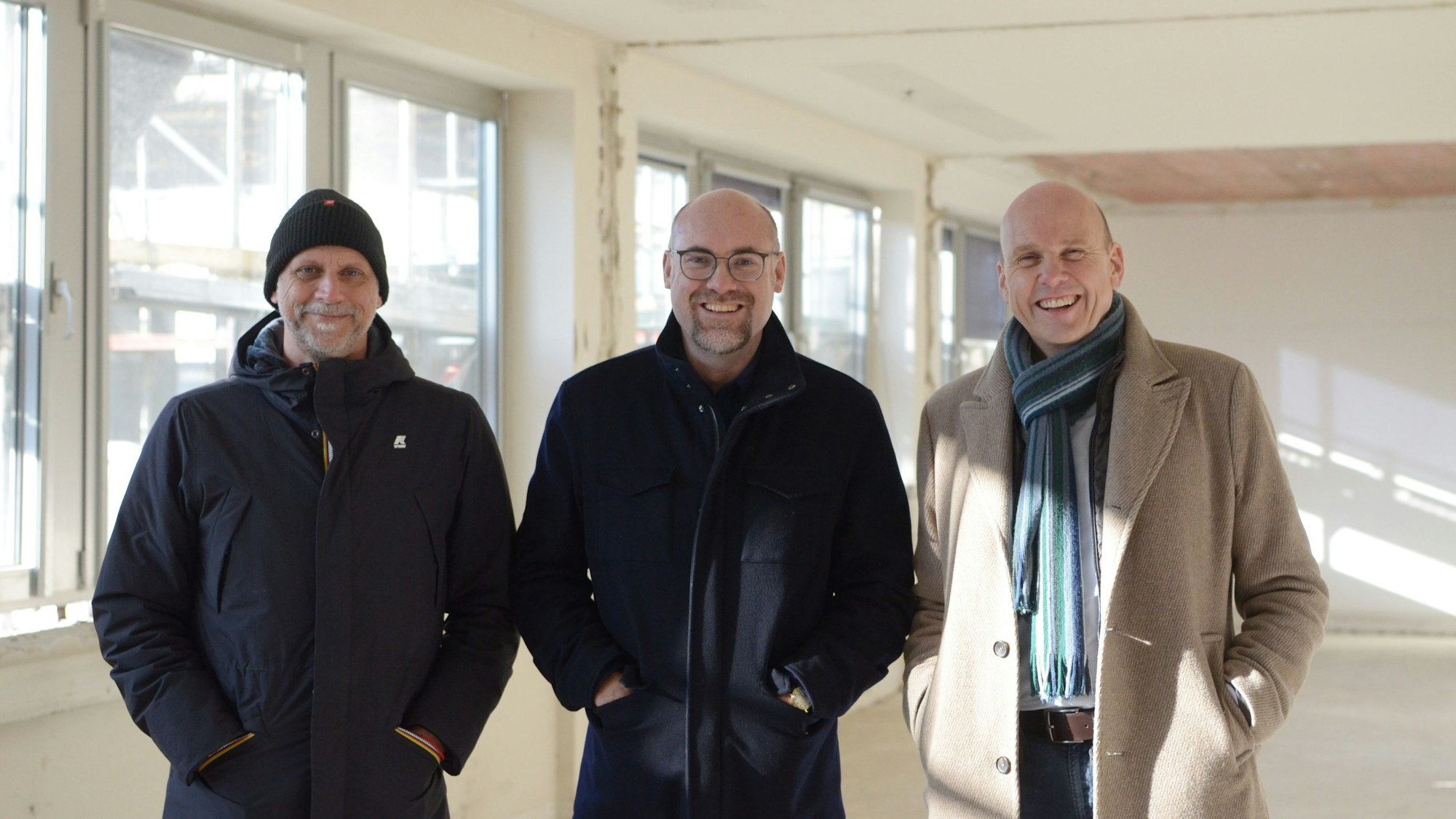 Drei Männer stehen nebeneinander in einem entkernten, lichtdurchfluteten Raum.