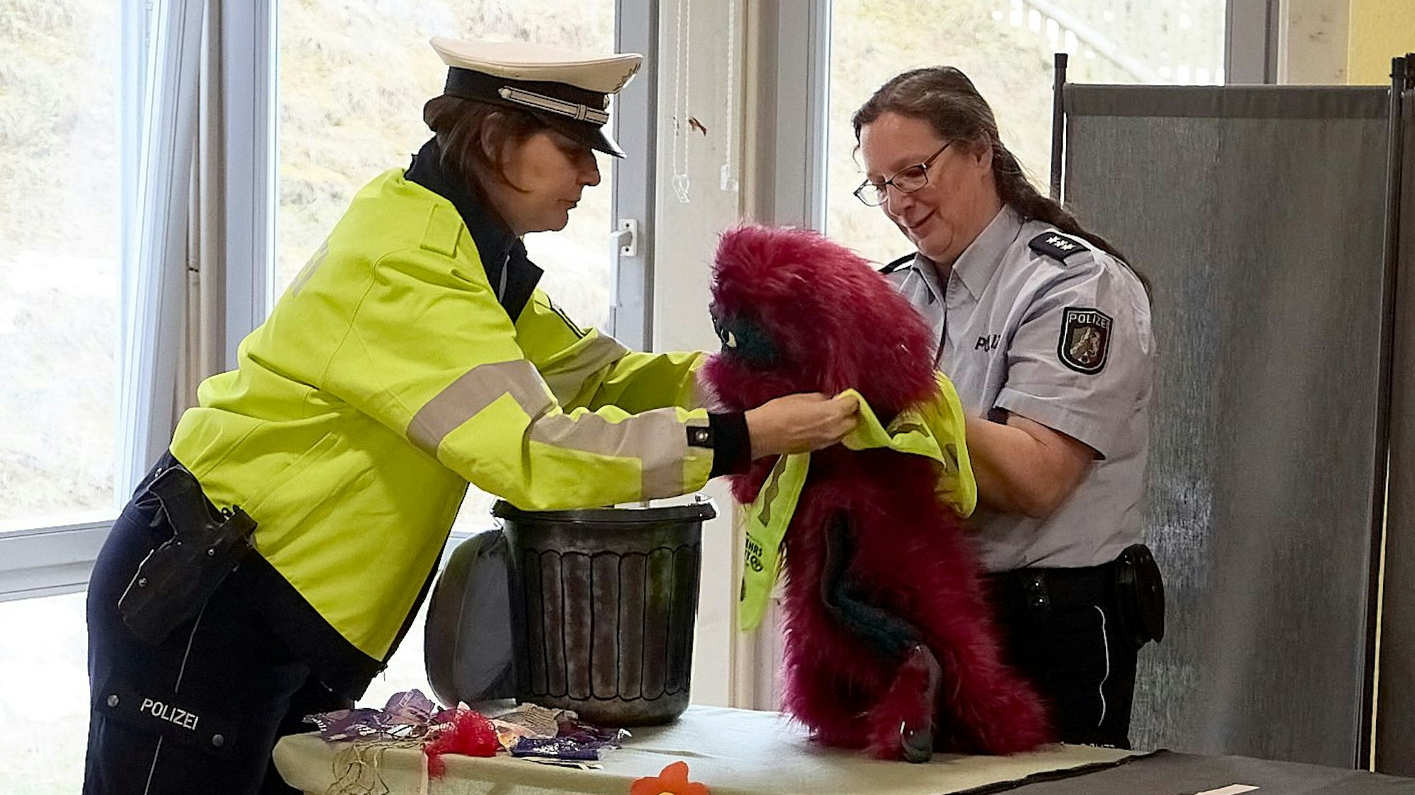 Zwei Polizistinnen in Uniform ziehen einer roten Plüschfigur eine gelbe Warnweste an. Die Figur ist das Müllmonster, das zu einem Theaterstück gehört, das die Polizei in einem Präventionsprogramm in Kindergärten einsetzt.