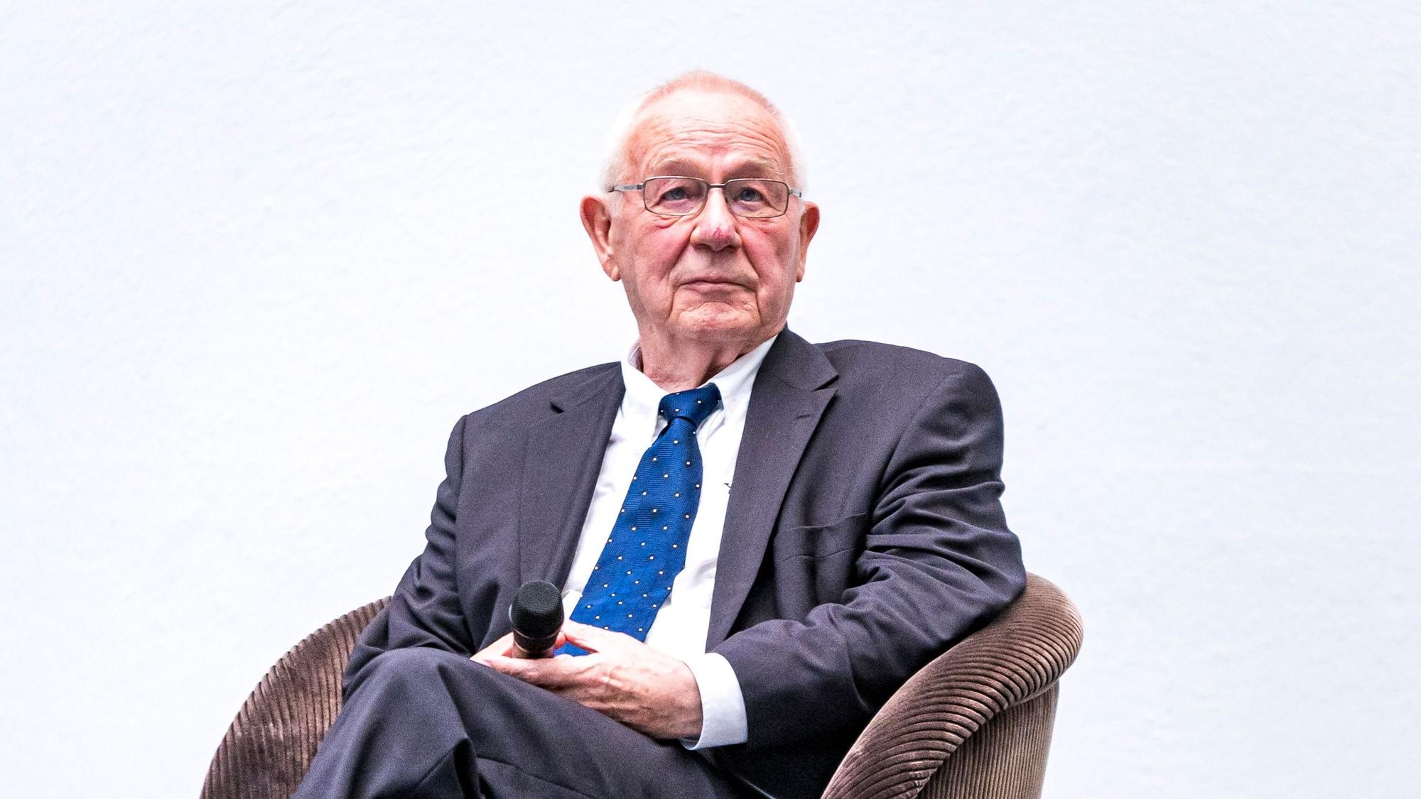 Armin Maiwald sitzt im Anzug und mit Mikrofon auf einer Bühne.