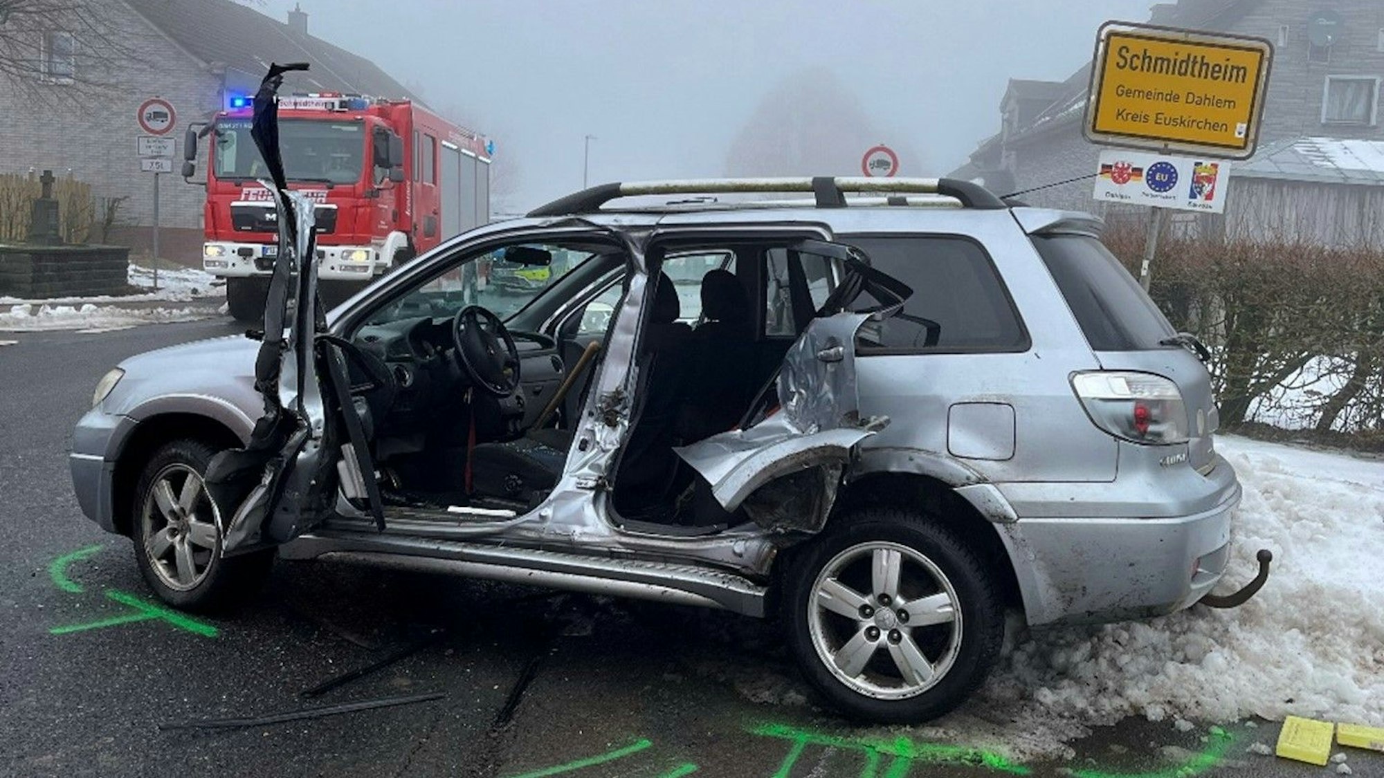 Ein silberfarbenes Auto steht nach einem Unfall massiv beschädigt auf einer Straße. Rechts dahinter ist das Ortsschild von Schmidtheim zu sehen, links ein Feuerwehrfahrzeug.