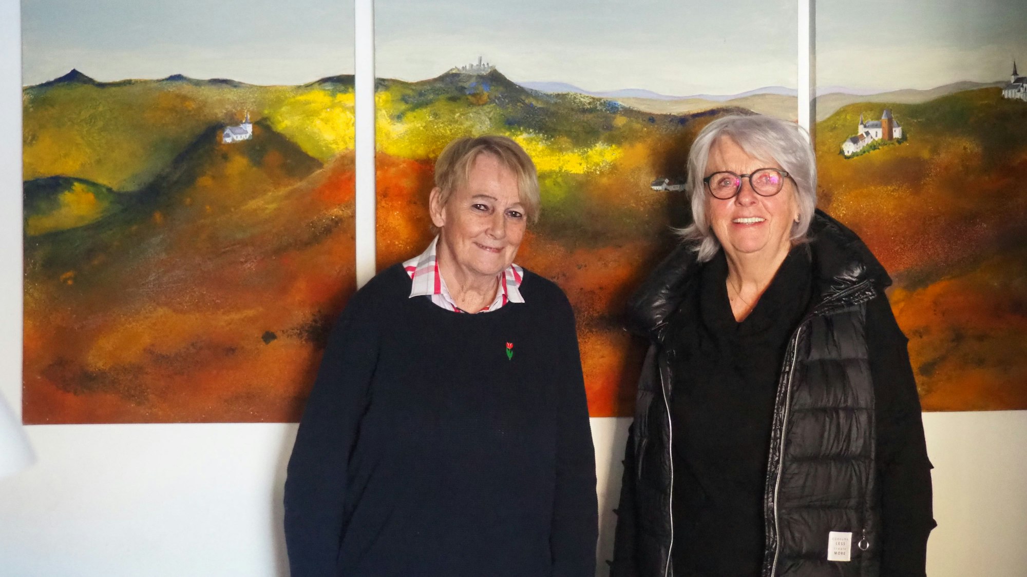 Zwei Frauen, Ulrike Ahrens und Gabriele Franzen, stehen vor einem dreiteiligen Gemälde in Grün- und Brauntönen.