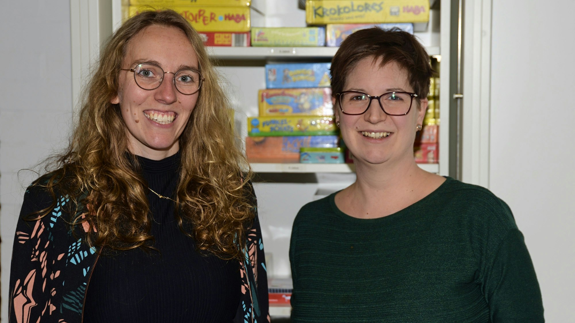 Zwei Frauen stehen in einer Bücherei und lächeln in die Kamera.