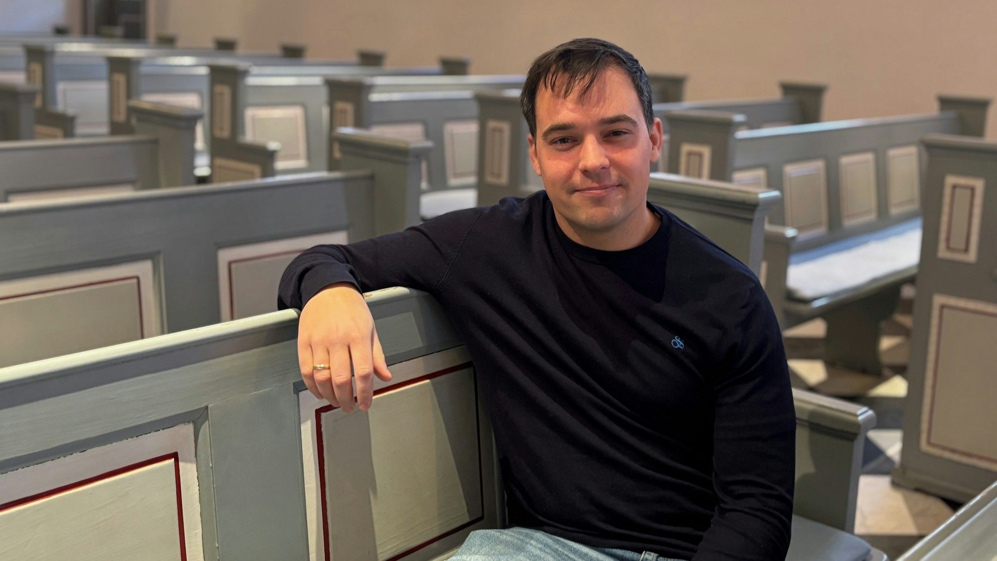 Auf dem Bild ist der neue Pfarrer in Frechen zu sehen, der auf einer Kirchenbank sitzt.