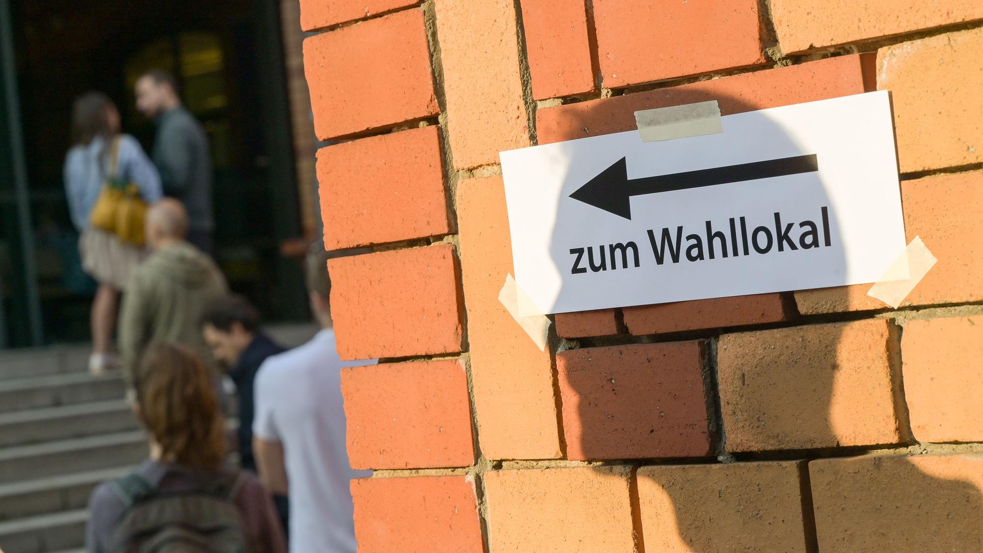 Wählerinnen und Wähler warten vor einem Wahllokal, während auf einer Mauer davor der Schatten eines Mannes auf einen Zettel mit der Aufschrift „Zum Wahllokal“ fällt.