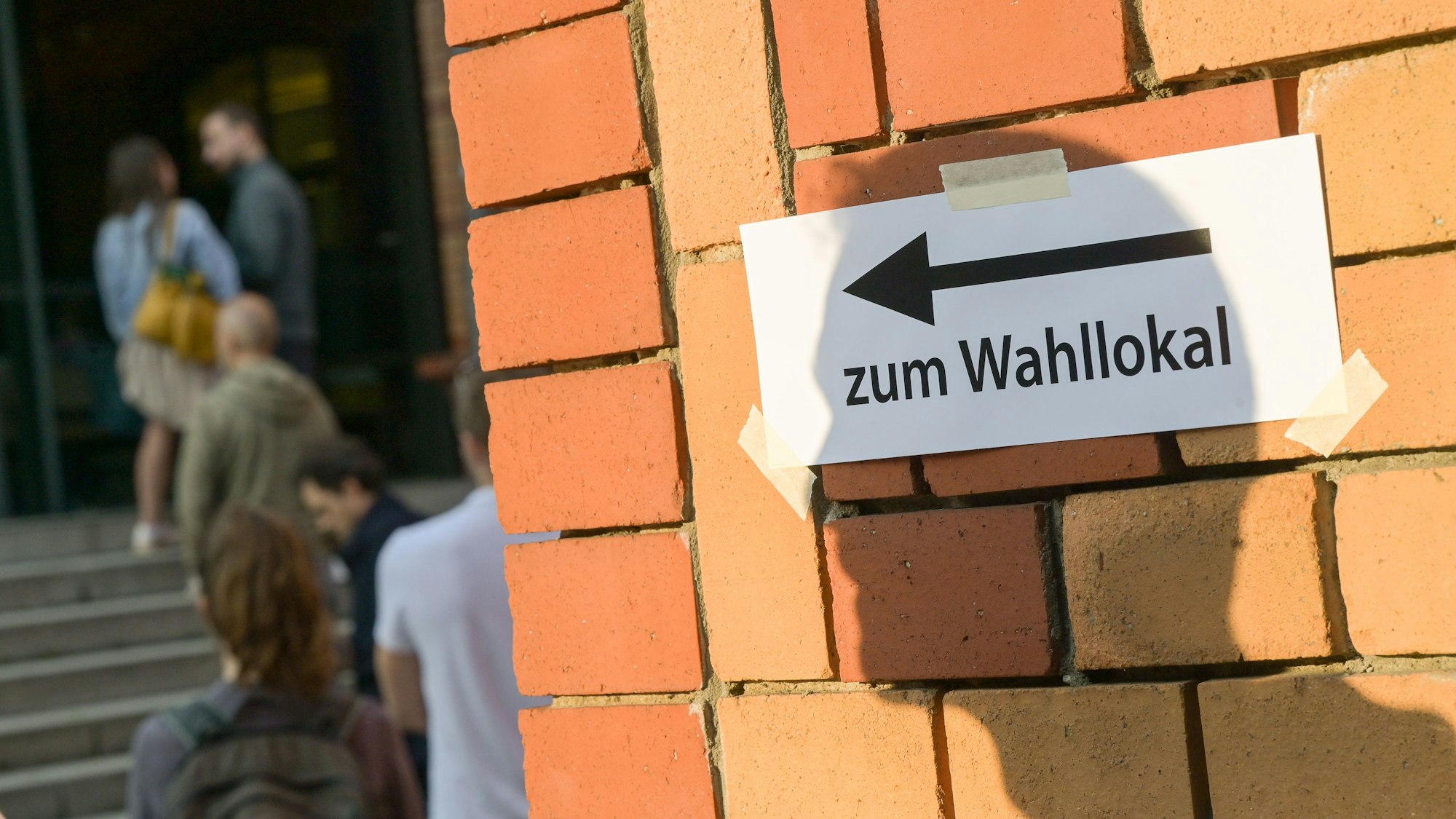 Wählerinnen und Wähler warten vor einem Wahllokal, während auf einer Mauer davor der Schatten eines Mannes auf einen Zettel mit der Aufschrift „Zum Wahllokal“ fällt.