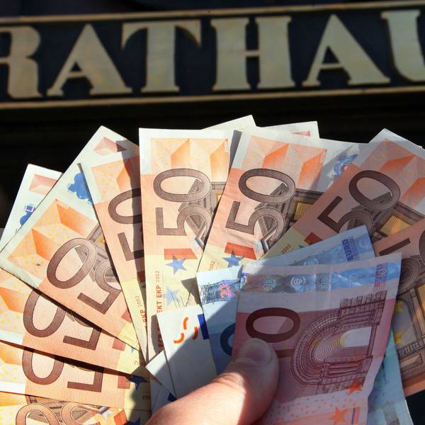 Eine Hand hält Geldscheine, vor allem Fünfzig-Euro-Scheine, vor dem Eingang zu einem Rathaus.