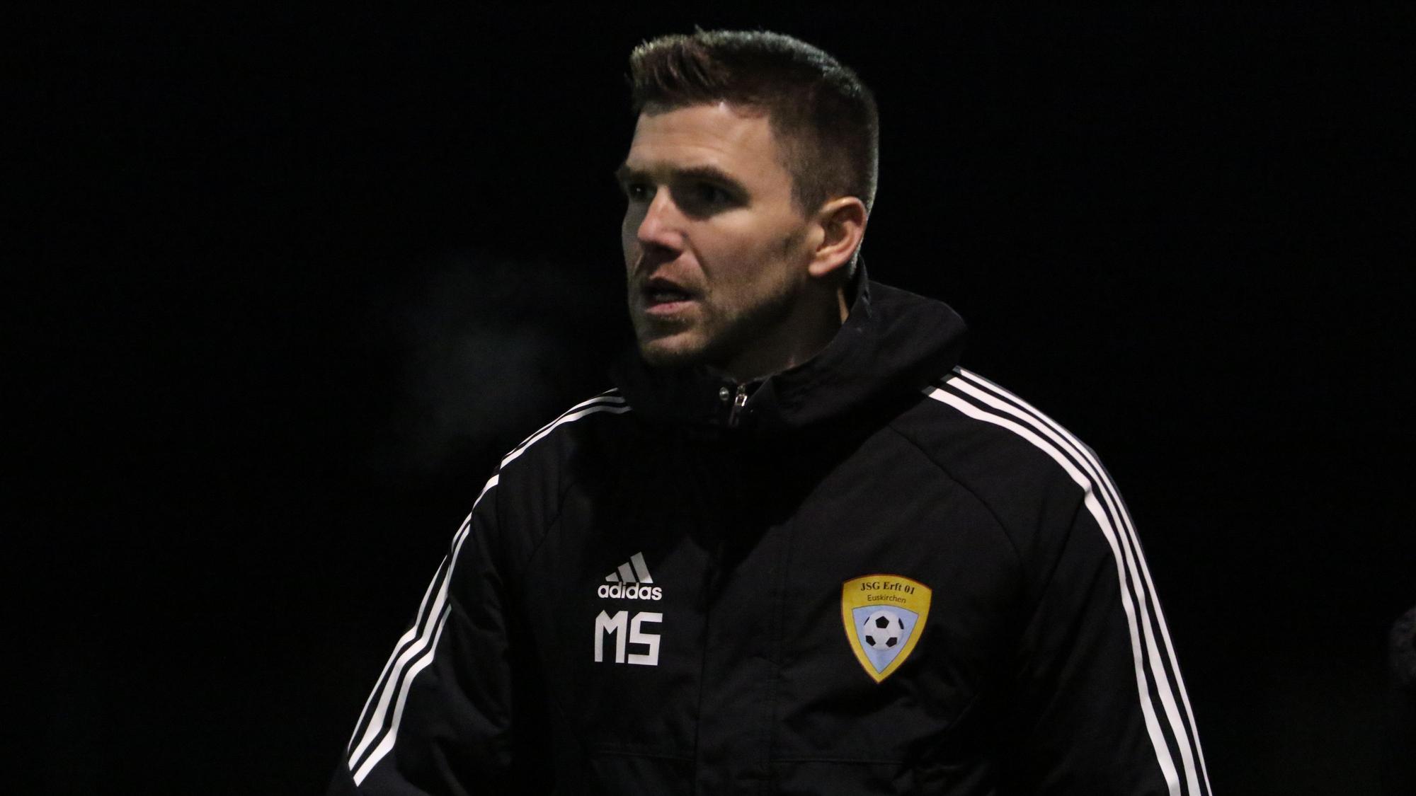 Der Trainer der A-Jugend der JSG Erft 01 Euskirchen, Marcel Schmitz, trägt eine dicke Jacke mit Initialien und Vereinslogo.