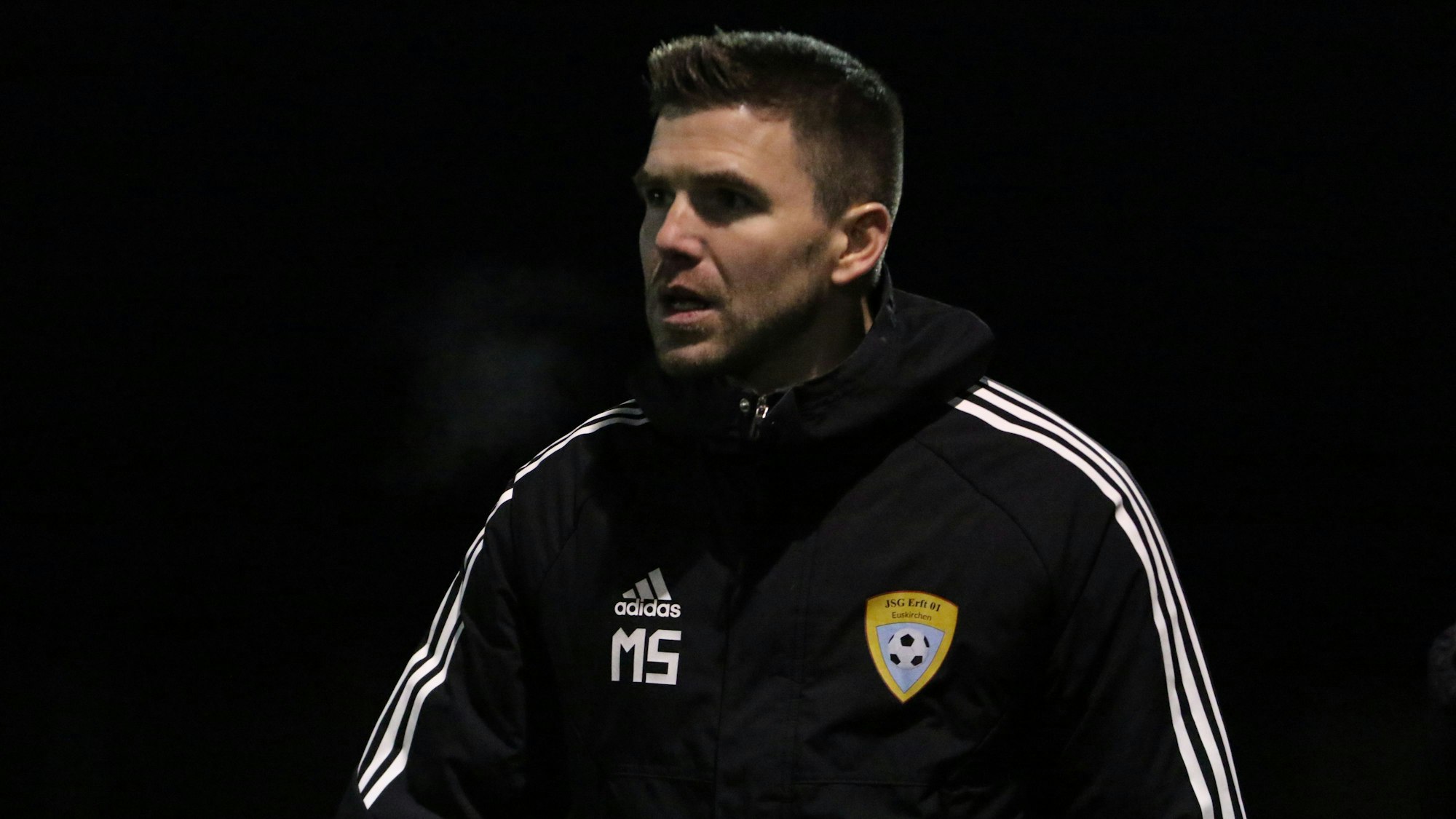 Der Trainer der A-Jugend der JSG Erft 01 Euskirchen, Marcel Schmitz, trägt eine dicke Jacke mit Initialien und Vereinslogo.