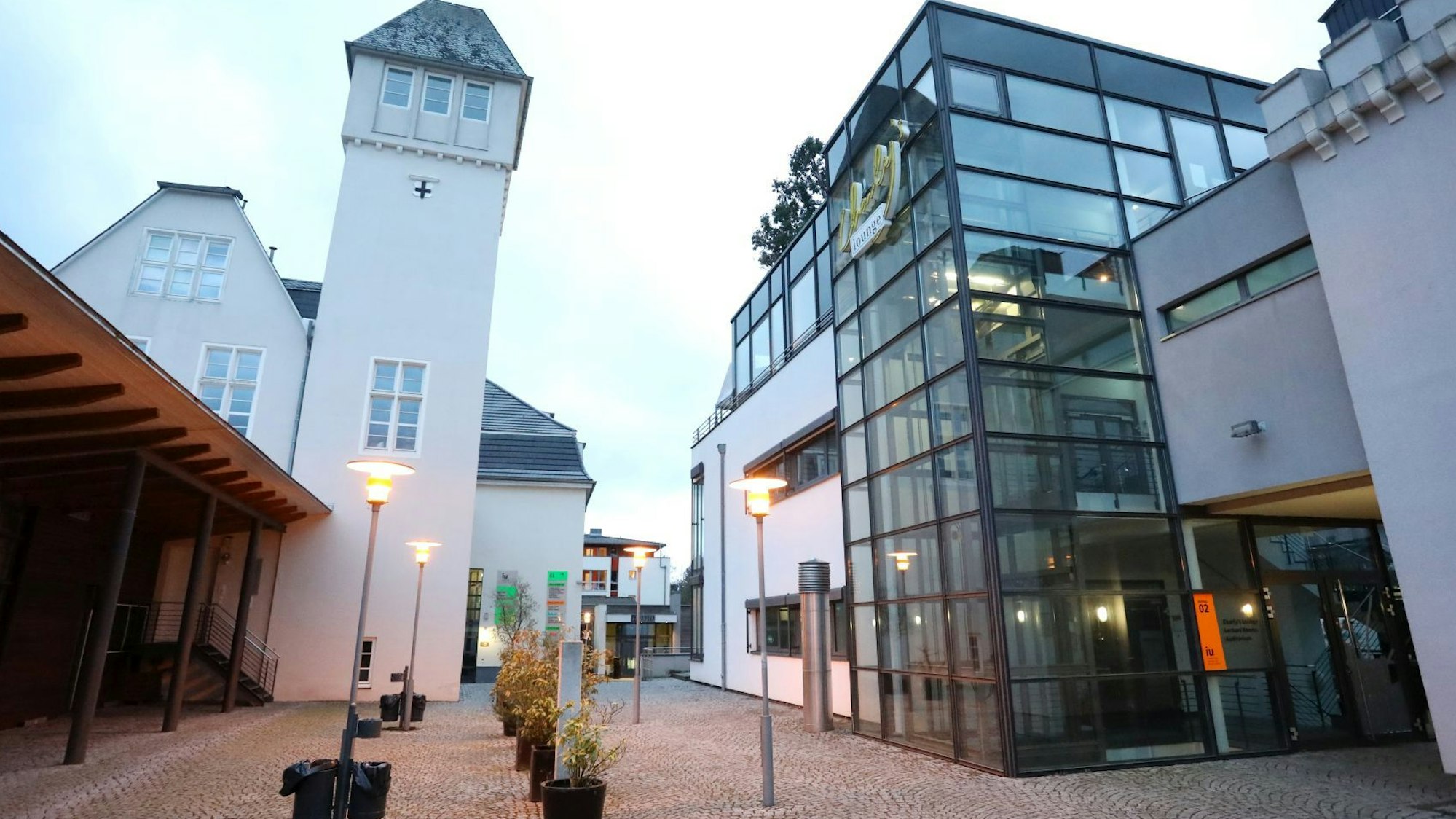 Ein historisches Gebäude mit einem hohen Turm neben einem Neubau mit Glasfassade. Auf dem Platz dazwischen stehen Straßenlaternen.