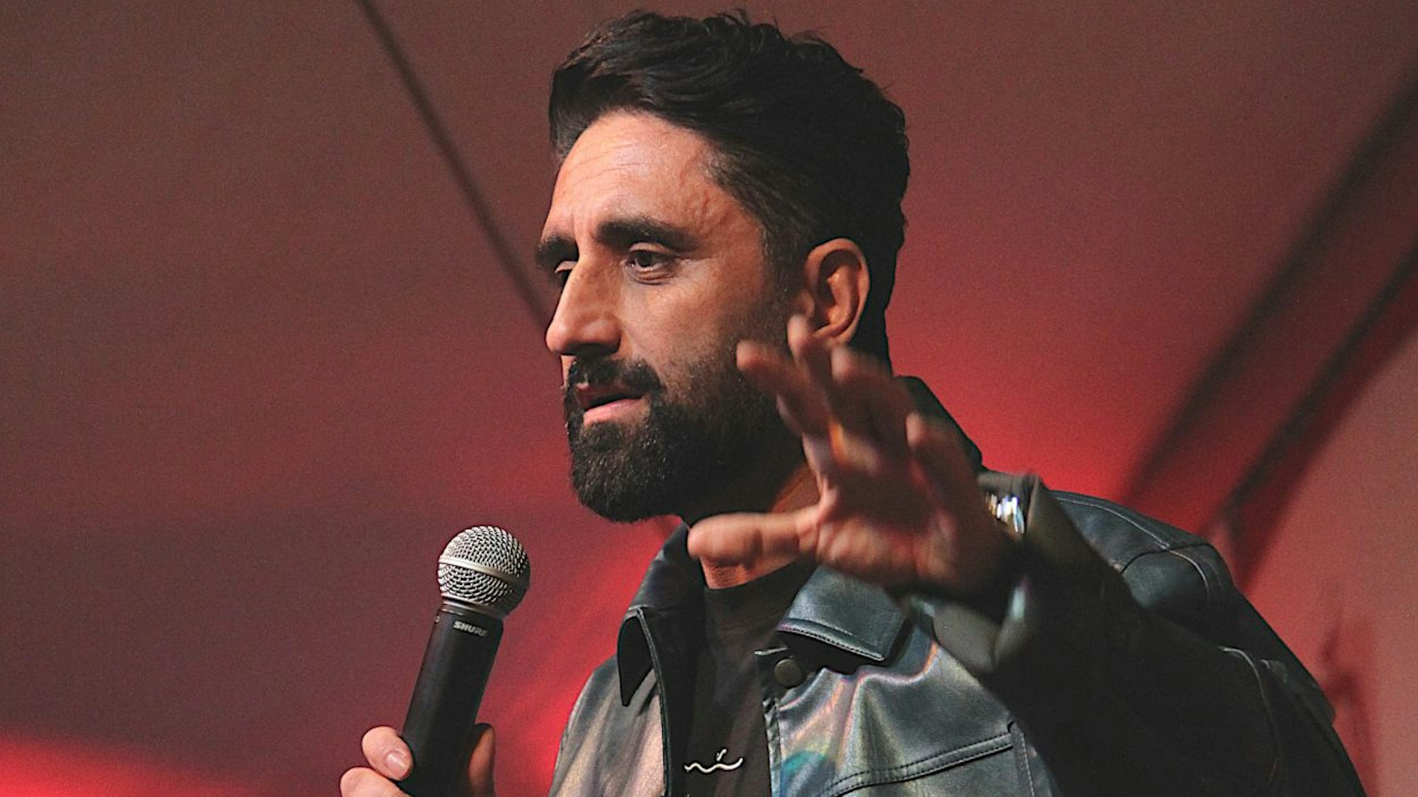 Comedian Djavid trägt eine schwarze Lederjacke und spricht in ein Mikrofon.