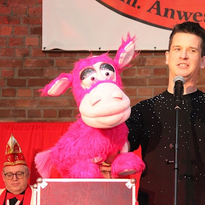 Bauchredner Tim Becker steht auf der Bühne in Mechernich-Vussem und spricht in ein Mikrofon. In der rechten hält er eine pinkfarbene Plüsch-Kuh.