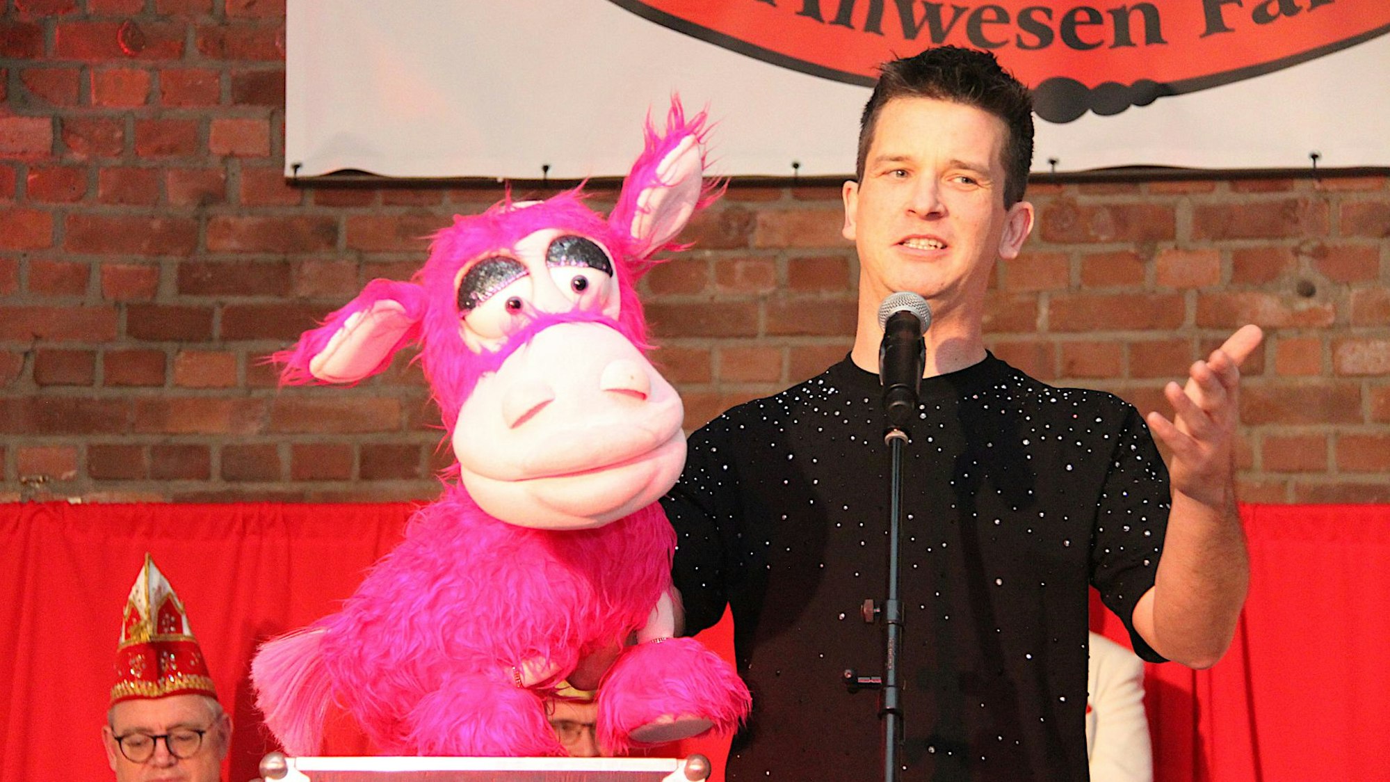 Bauchredner Tim Becker steht auf der Bühne in Mechernich-Vussem und spricht in ein Mikrofon. In der rechten hält er eine pinkfarbene Plüsch-Kuh.