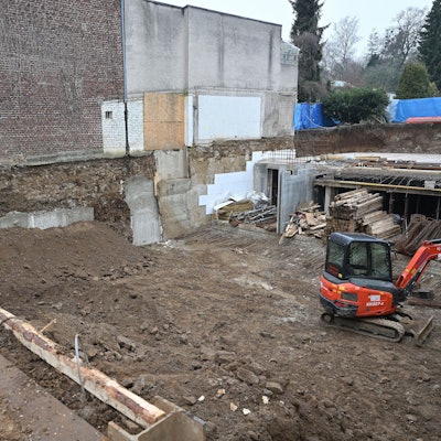 Blick von oben in die Baugrube. Man sieht einen Erdhaufen vor der Hauswand, Baumaterial sowie einen Bagger.