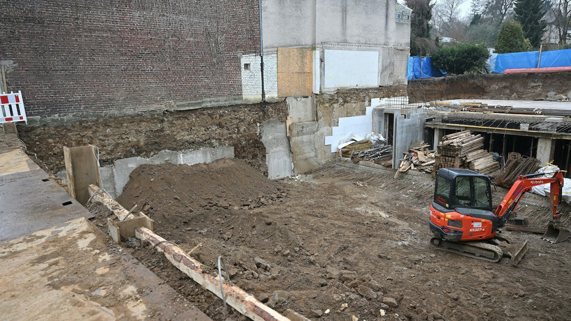 Blick von oben in die Baugrube. Man sieht einen Erdhaufen vor der Hauswand, Baumaterial sowie einen Bagger.