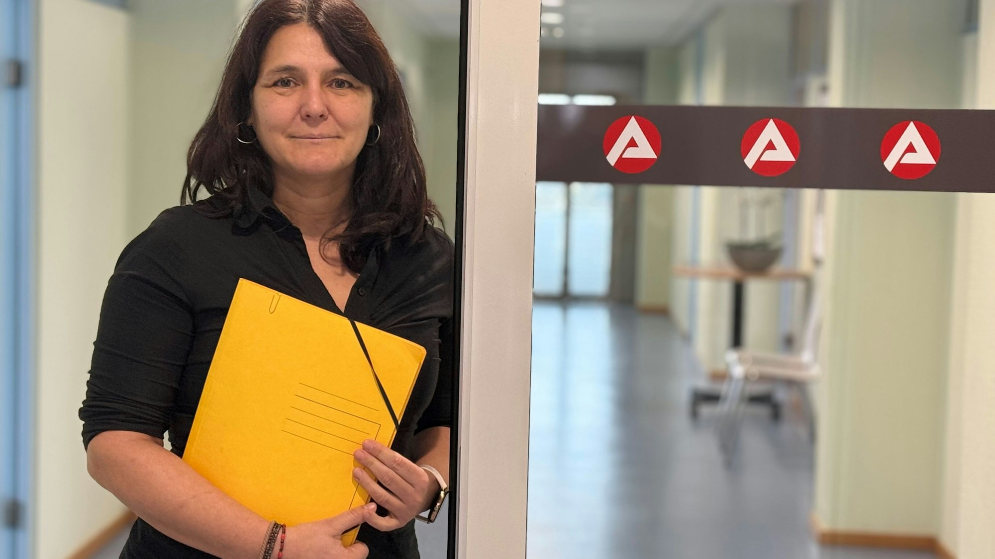 Anja Daub ist Geschäftsführerin operativ der Agentur für Arbeit in Brühl. Sie hält eine gelbe Aktenmappe in den Händen und steht an einer Glastür mit drei Logos der Arbeitsagentur.