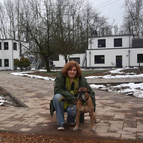 Zu sehen ist eine Frau mit einem alten Boxerhund vor neuen Gebäuden.