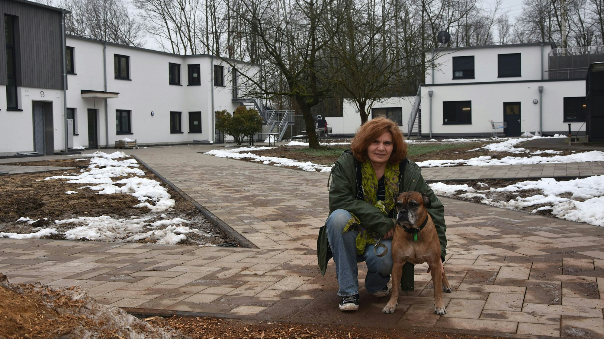 Zu sehen ist eine Frau mit einem alten Boxerhund vor neuen Gebäuden.
