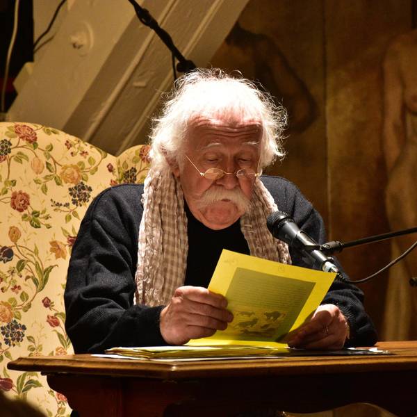 Ein Mann mit weißen Haaren und einem breiten weißen Schnurrbart rezitiert an einem Tisch.