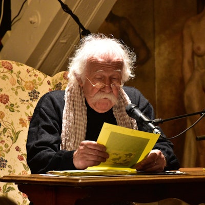 Ein Mann mit weißen Haaren und einem breiten weißen Schnurrbart rezitiert an einem Tisch.