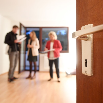 Eine Maklerin spricht mit Interessenten bei einer Wohnungsbesichtigung, während im Vordergrund eine Türklinke zu sehen ist.