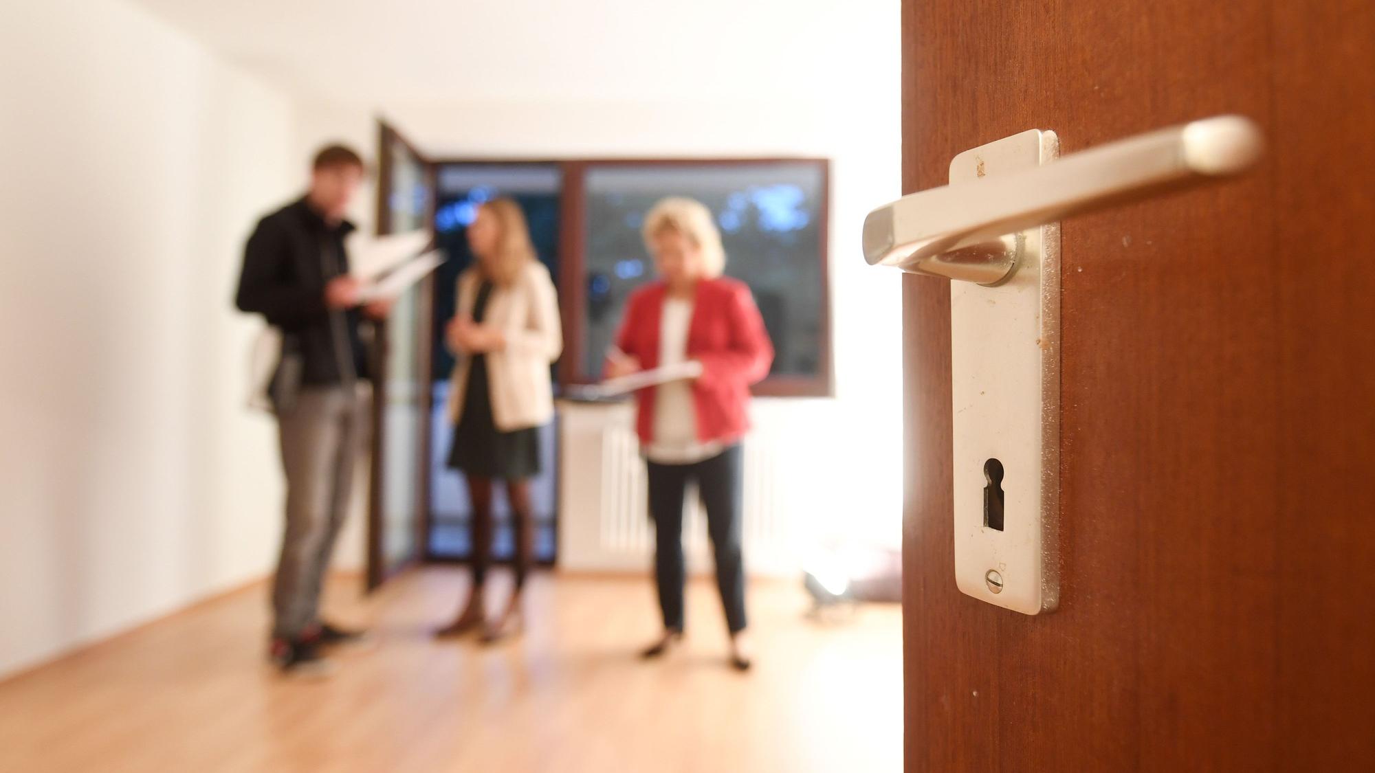 Eine Maklerin spricht mit Interessenten bei einer Wohnungsbesichtigung, während im Vordergrund eine Türklinke zu sehen ist.