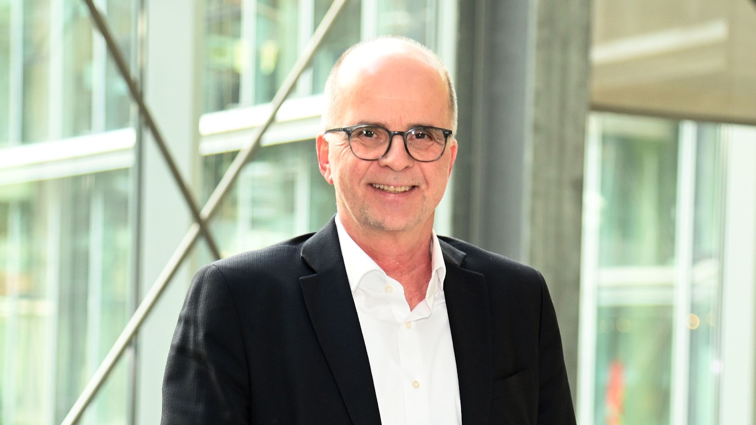 Ein Mann mit wenig Haaren und Brille steht lachend vor einer Glasfront.