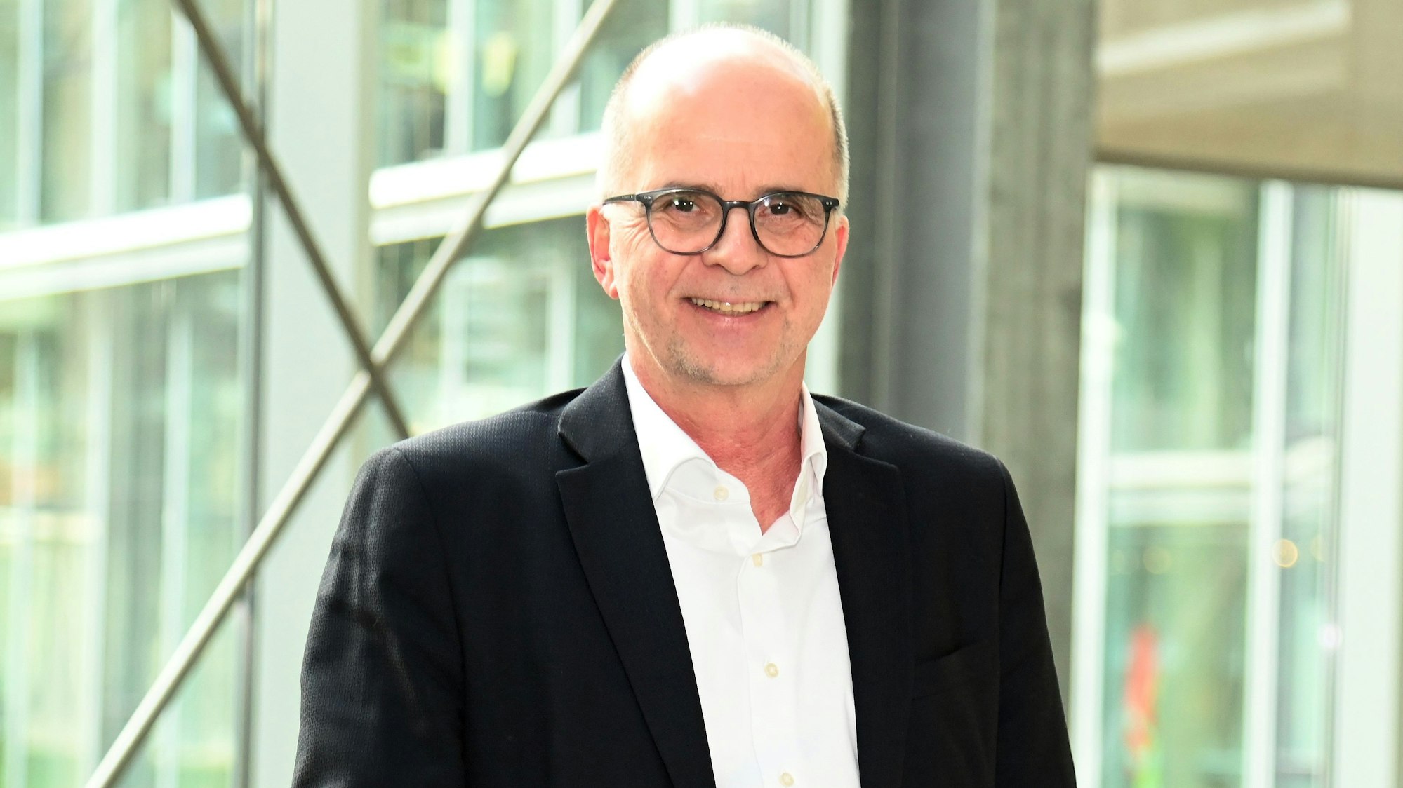 Ein Mann mit wenig Haaren und Brille steht lachend vor einer Glasfront.