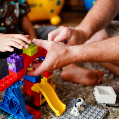 Das Bild zeigt Männerhände, die mit einem Kind einen Turm aus Klemmbausteinen bauen.