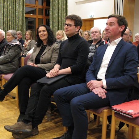 Zwei Männer und eine Frau sitzen in der erste Reihe in einem Saal.