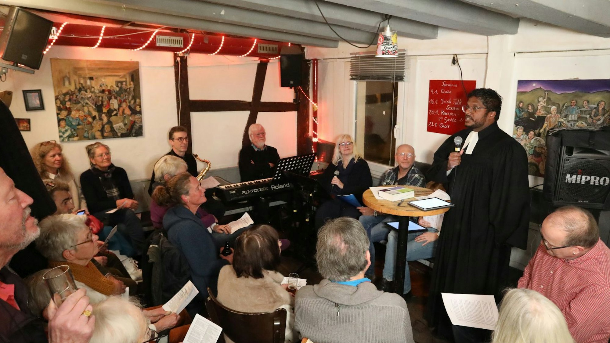 Mehrere Menschen in einem kleinen Raum, der Diakon hält ein Mikrofon in der Hand, link sitzen Musiker.