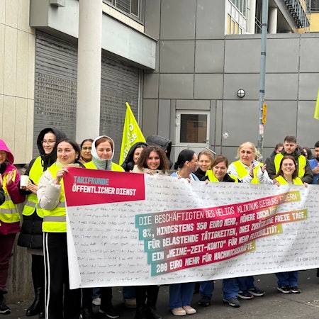 Pflegekräfte stehen vor dem Helios Krankenhaus in Siegburg und halten ein Banner. Darauf steht „Tarifrunde Öffentlicher Dienst“.