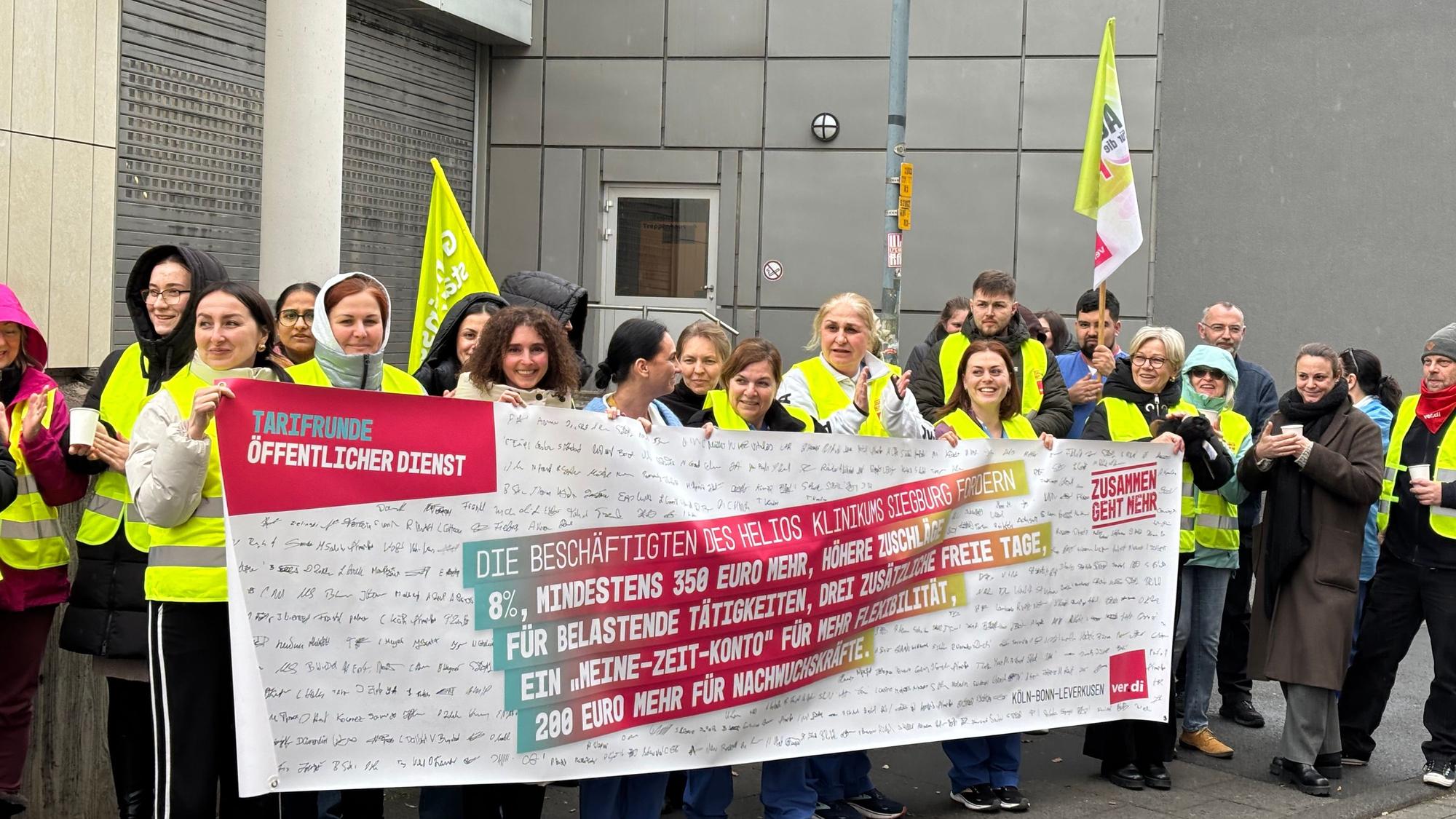 Pflegekräfte stehen vor dem Helios Krankenhaus in Siegburg und halten ein Banner. Darauf steht „Tarifrunde Öffentlicher Dienst“.