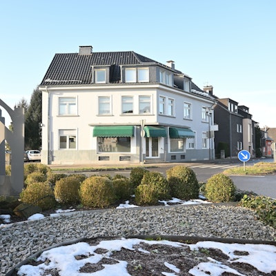 Ein Kreisverkehr mit bepflanzter Mittelinsel, dahinter ein Haus mit Ladenlokal.