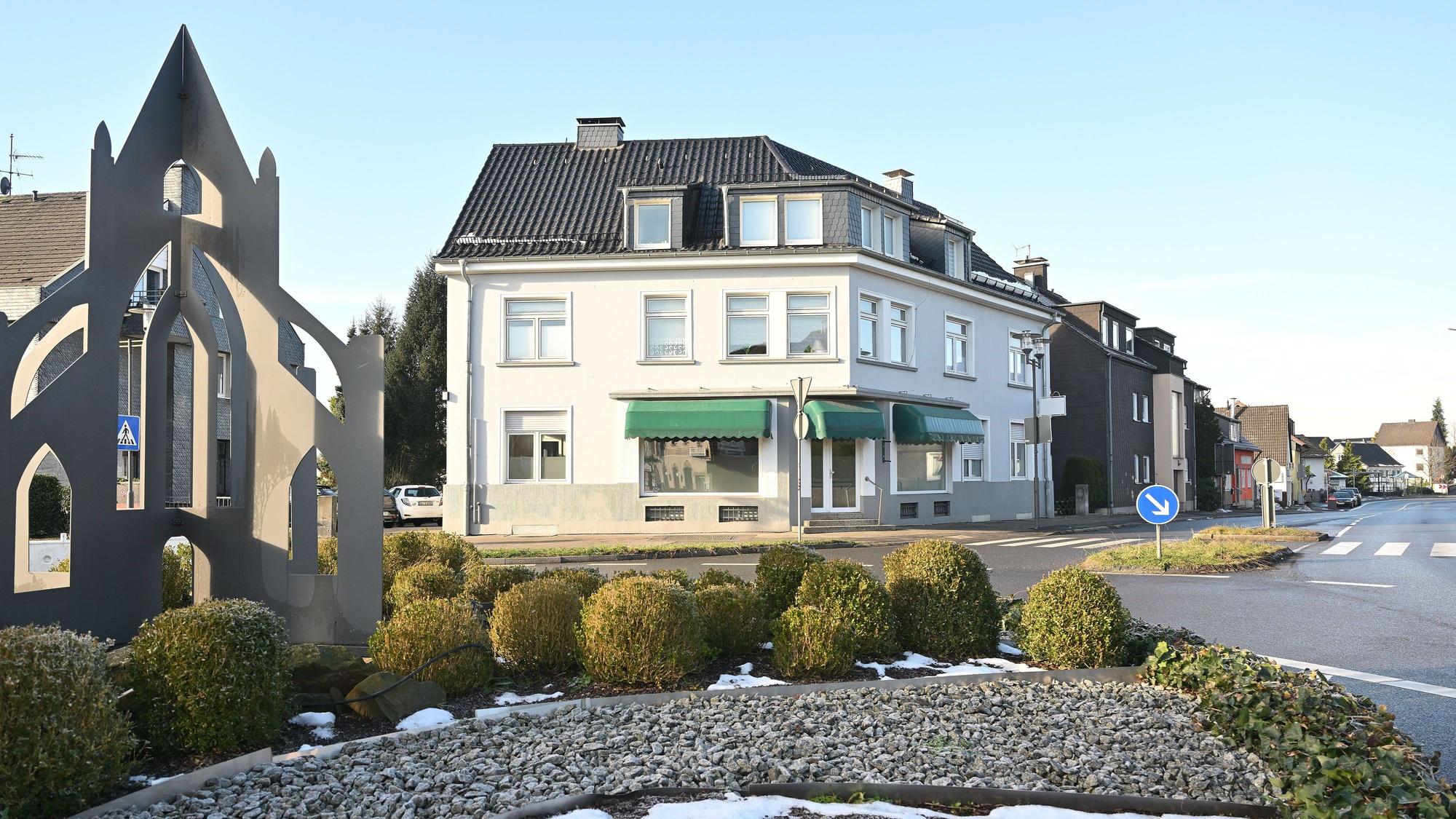Ein Kreisverkehr mit bepflanzter Mittelinsel, dahinter ein Haus mit Ladenlokal.