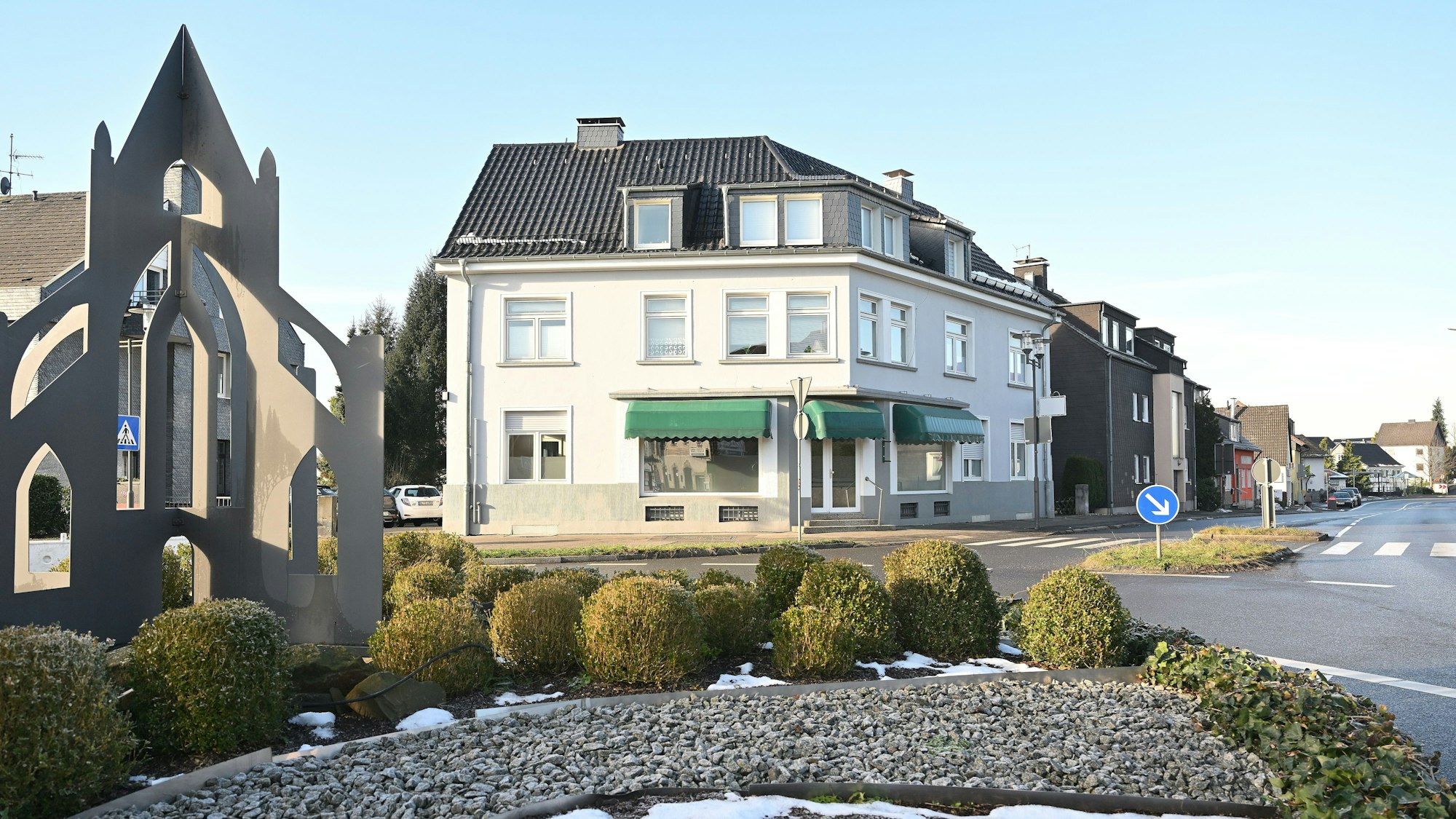 Ein Kreisverkehr mit bepflanzter Mittelinsel, dahinter ein Haus mit Ladenlokal.