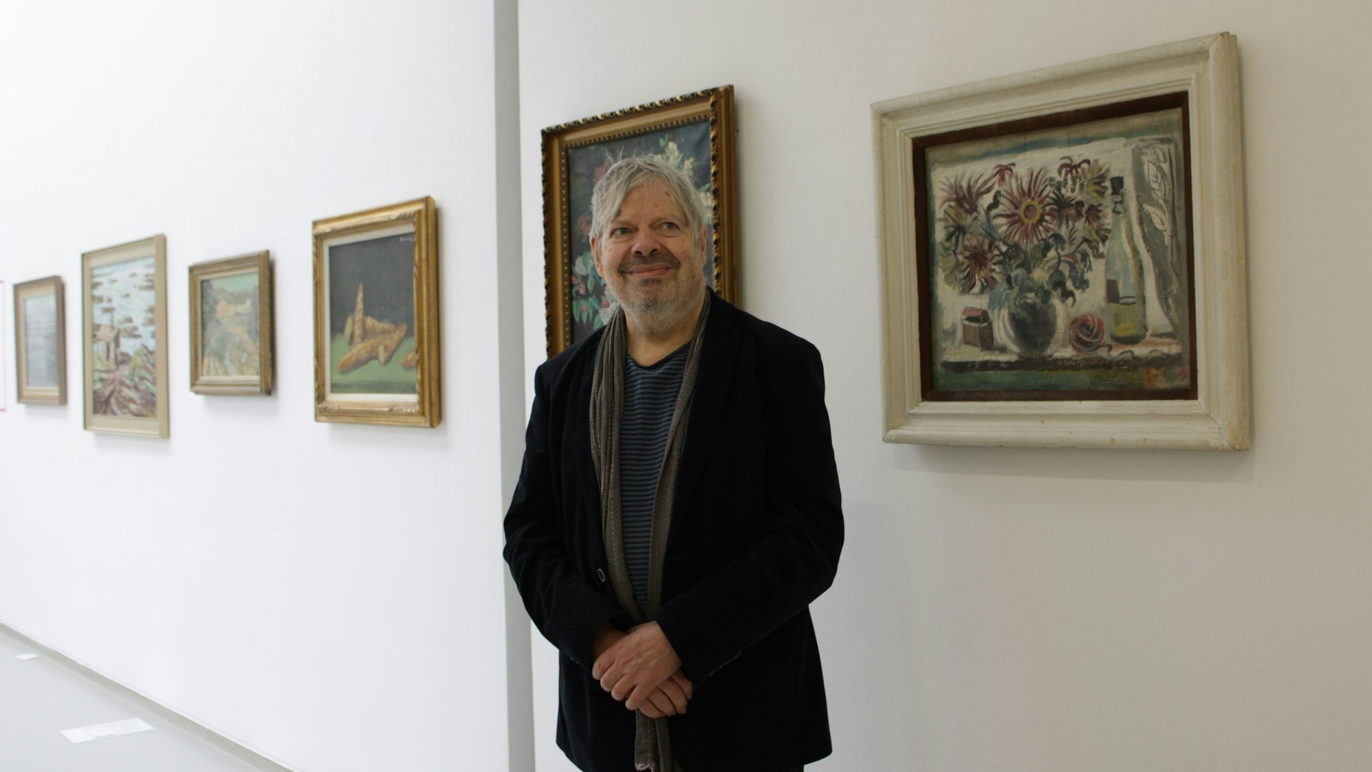 Ein Mann steht in einer Ausstellung, die Hände gekreuzt, neben ihm ist ein Stillleben zu sehen, das eine Blumenvase, eine Flasche und ein Wollknäuel zeigt.