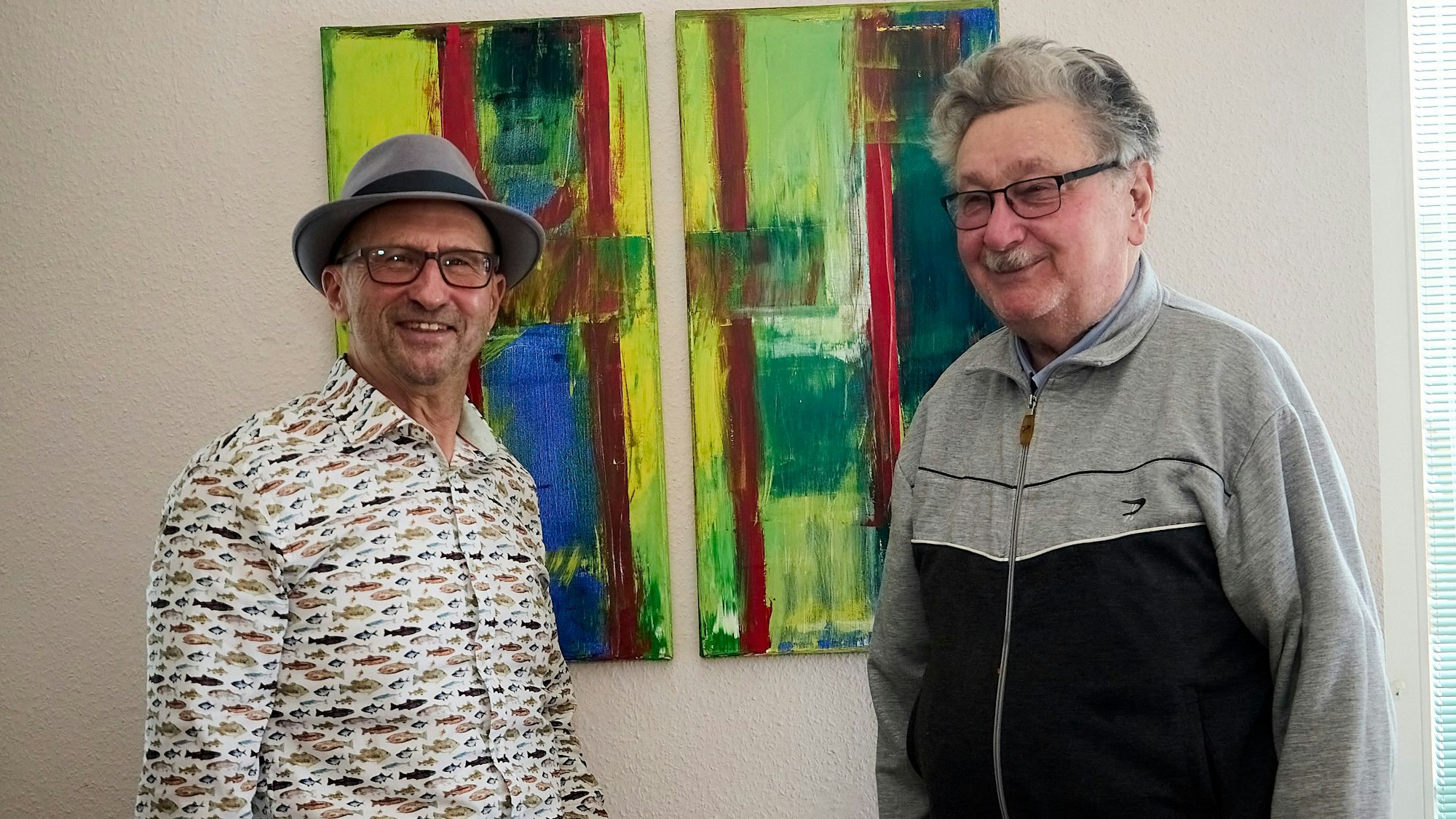 Zwei Männer, Heinz-Bert Weimbs und Albert Breuer, stehen vor einem bunten Gemälde, das an einer weißen Wand hängt.