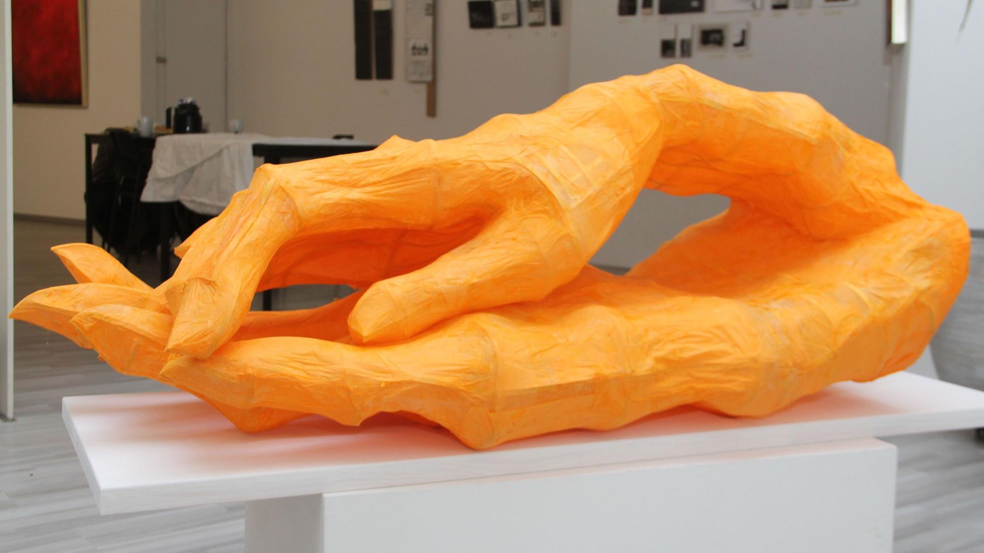 Eine orangefarbene Skulptur, die zwei zusammenhängende Hände darstellt, liegt auf einem Ausstellungstisch in einer Galerie.