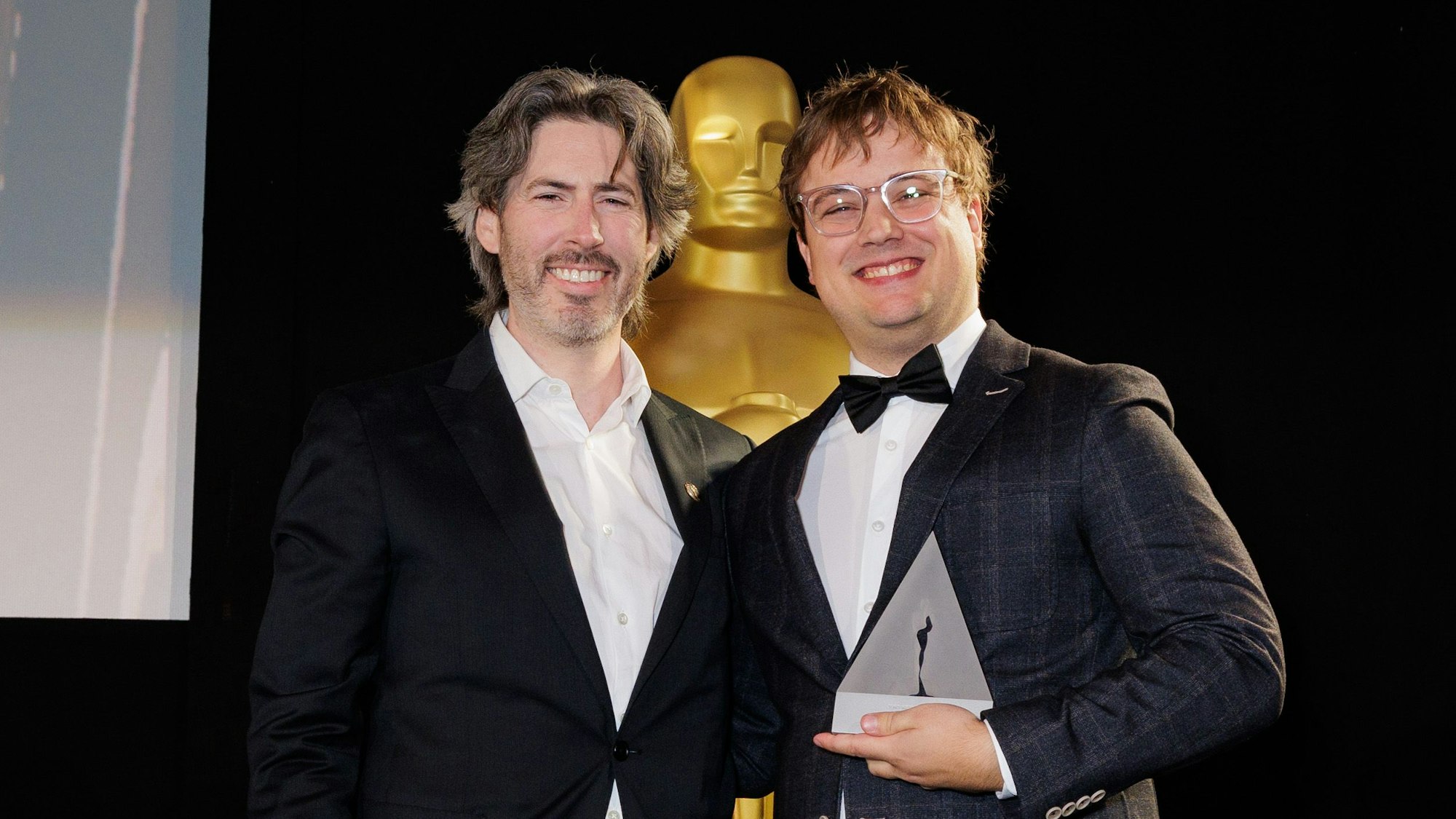 Zwei Männer vor einer großen Oscar-Statue.