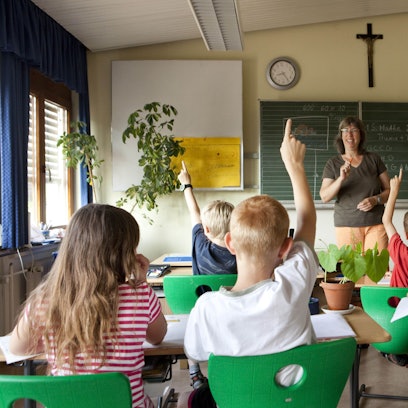Das Foto zeigt Schüler und eine Lehrerin in einem Klassenzimmer, an der Wand hängt ein Kreuz.