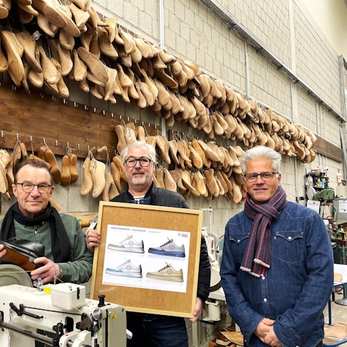 Drei Männer stehen in einer Werkstatt und halten ein Bild in den Händen, auf dem der neue Sneaker zu sehen ist. An der Wand hängen Leisten, Nachbildungen von original Füßen und im Vordergrund eine Nähmaschine.