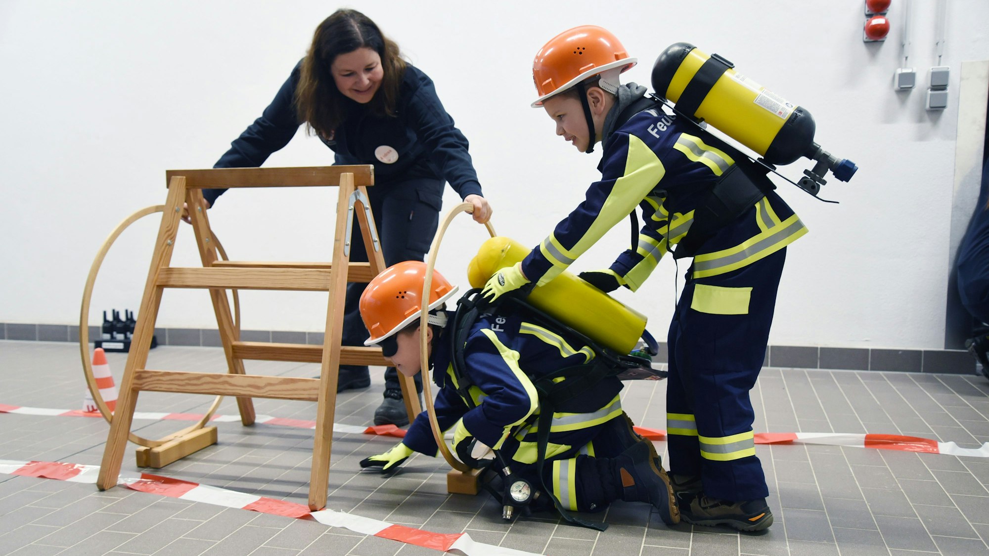 Zwei Kinder in Feuerwehruniform durchlaufen unter Anleitung einen Hindernisparcours.