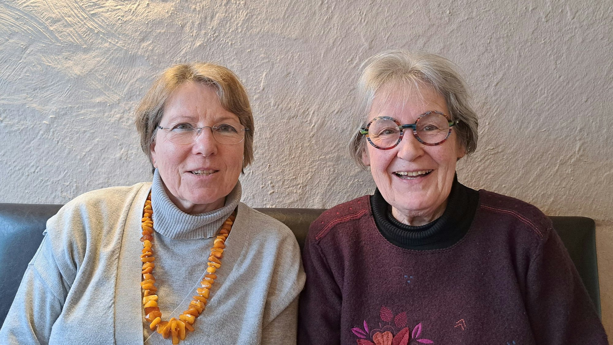 Zwei ältere Frauen sitzen auf einer schwarzen Couch und lächeln in die Kamera.