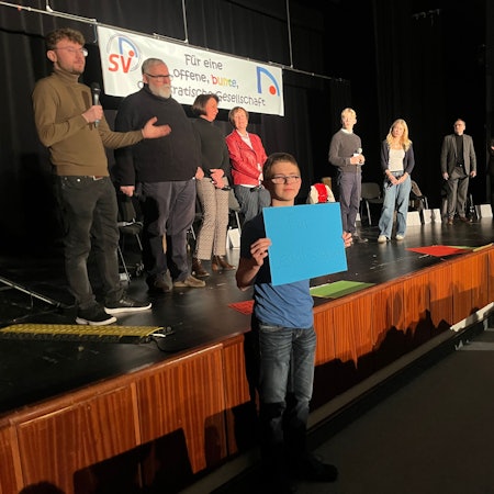 Sechs Politiker und zwei Schüler stehen auf einer Bühne, drei Schüler halten Plakate mit Fragen hoch.