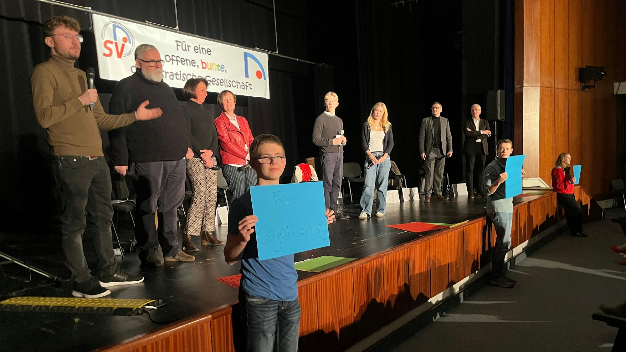 Sechs Politiker und zwei Schüler stehen auf einer Bühne, drei Schüler halten Plakate mit Fragen hoch.