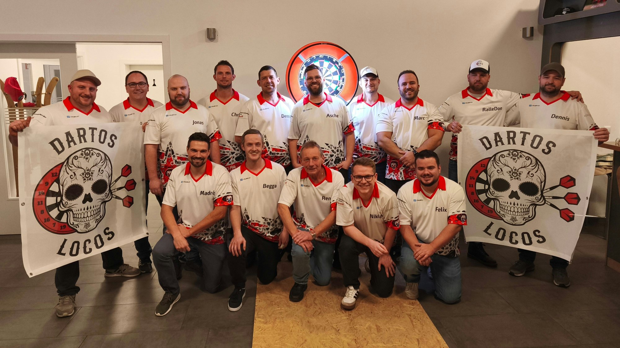 Die Spieler der Dartos Locos aus Billig posieren für die Kamera, links und rechts halten sie Fahnen mit dem Teamlogo hoch. Im Hintergrund ist eine beleuchtete Darts-Scheibe zu erkennen.